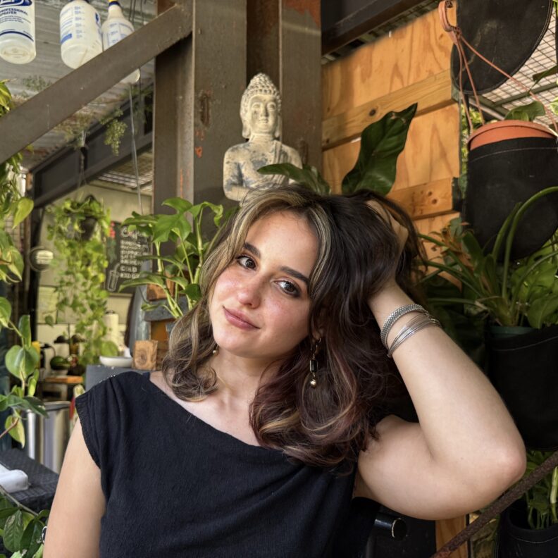 A woman posing with her hand in her hair in a room with multiple potted plants.