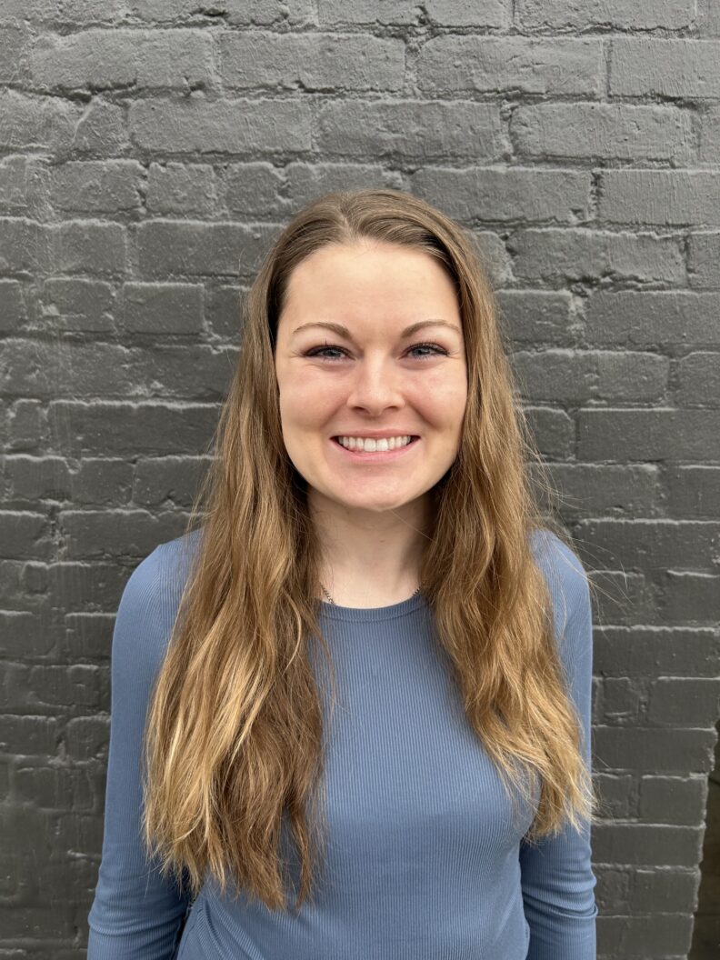 A woman in a blue shirt standing in front of a gray brick wall.