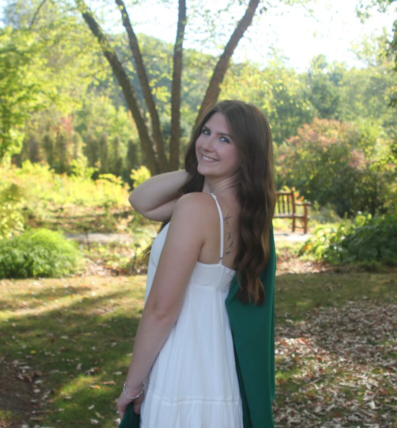 A woman looking over her shoulder standing in front of a green landscape.
