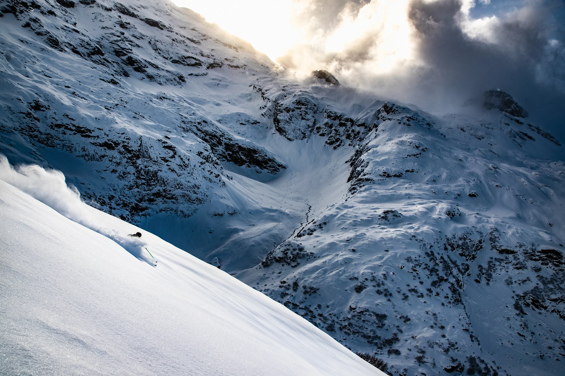 A breathtaking mountain edge covered in snow and large snow-dusted rocks. A skier in motion descends from the ridge, carving through the powder and sending a spray of snow into the sunlight as the sun breaks through the clouds