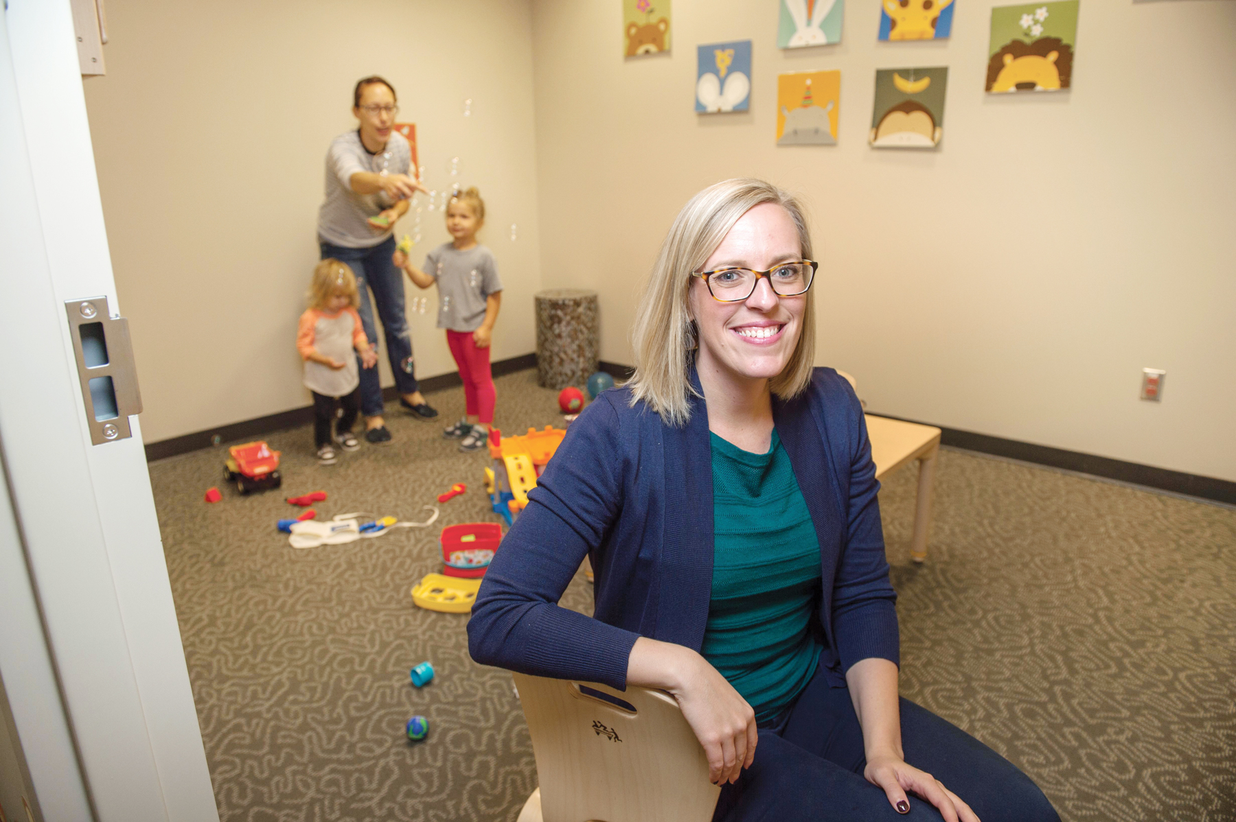 Professor Lauren Thompson is sitting in a chair, smiling with kids in the background, blowing bubbles.