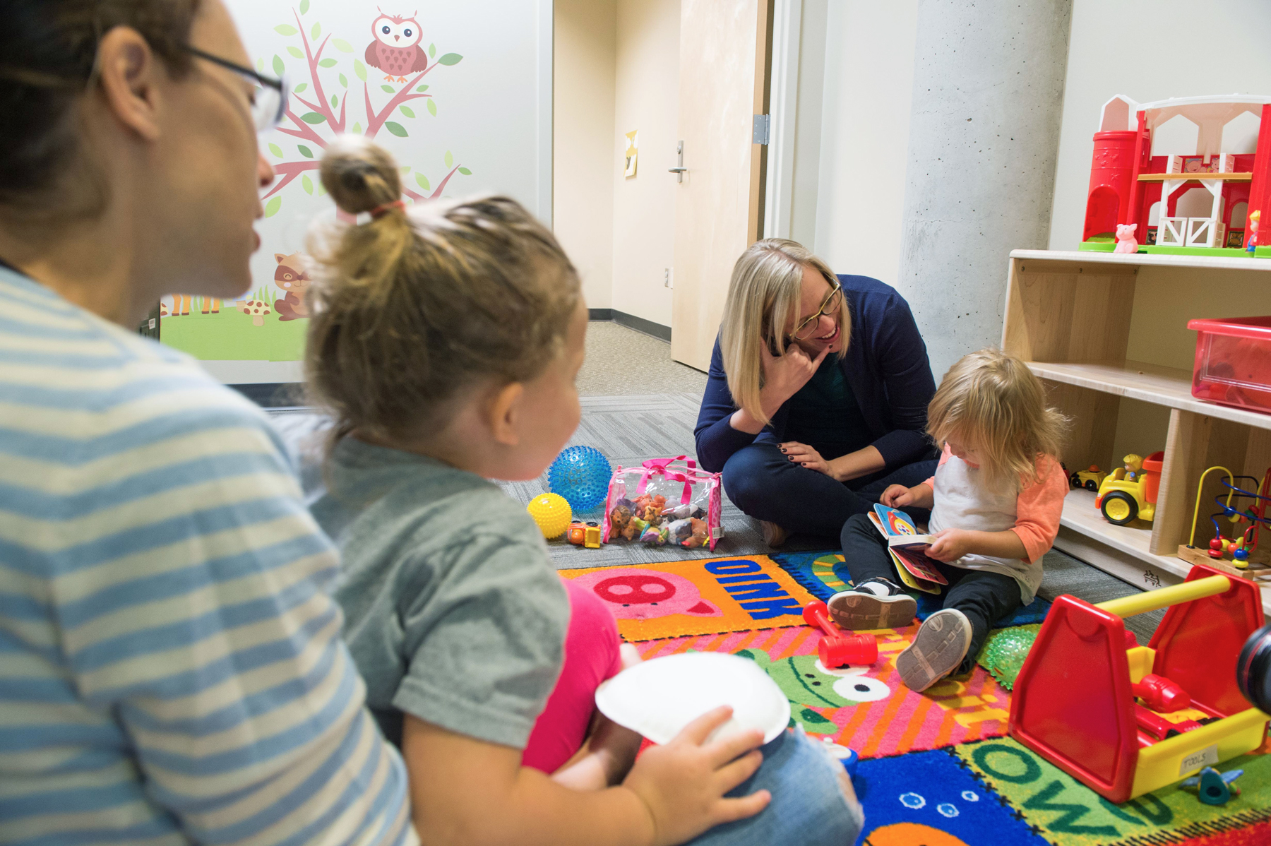 A professor sitting on the floor with a child looking at a book as another parent and child look on.