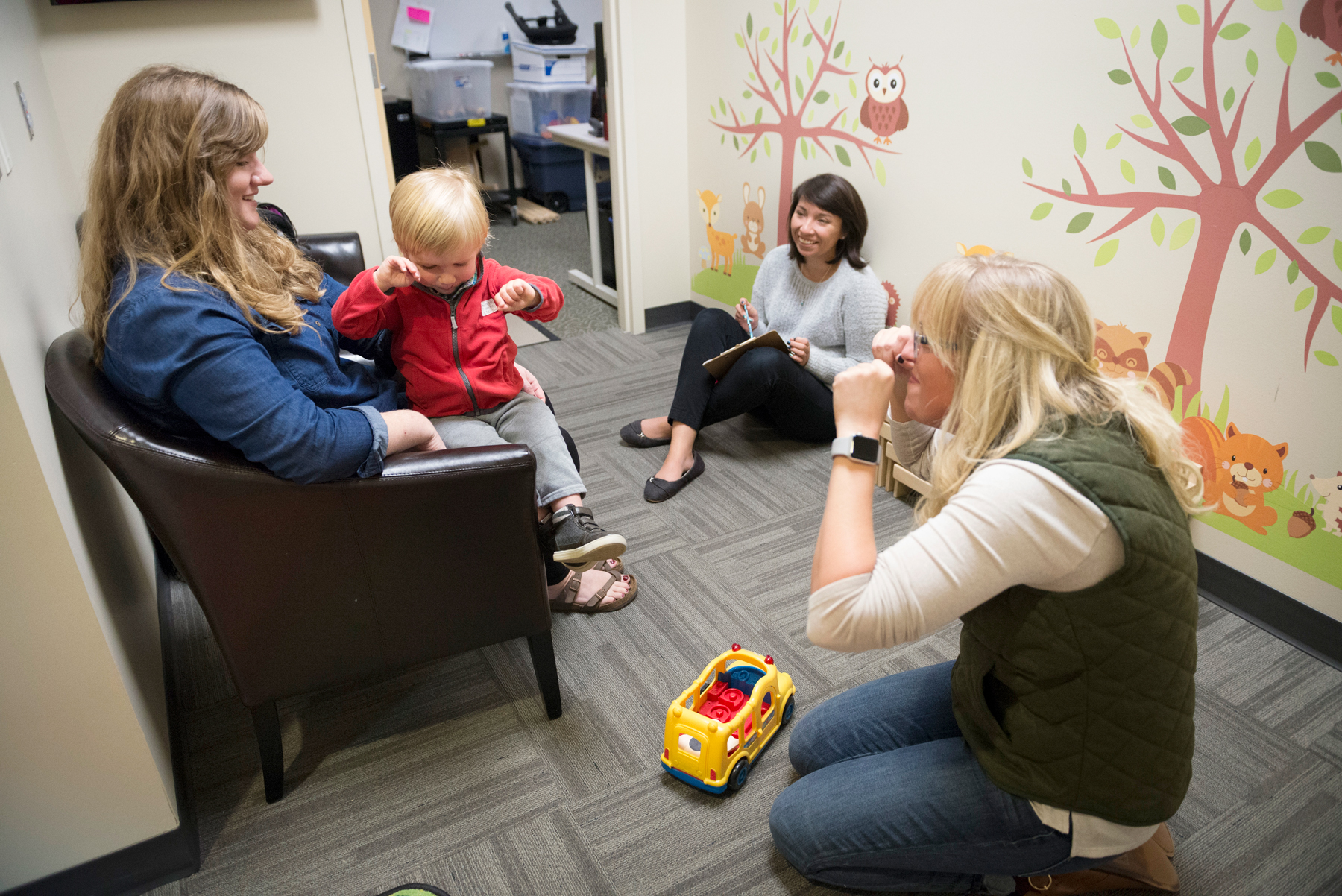 A professor sitting on floor in the play room with a parent and child rubbing their eyes.