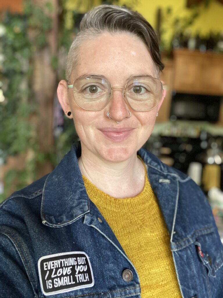 A person with short greyish hair wearing glasses, a yellow sweater, and a denim jacket smiling at the camera
