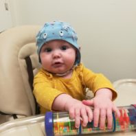 Baby boy in high chair wearing blue lab helmet and playing with toy