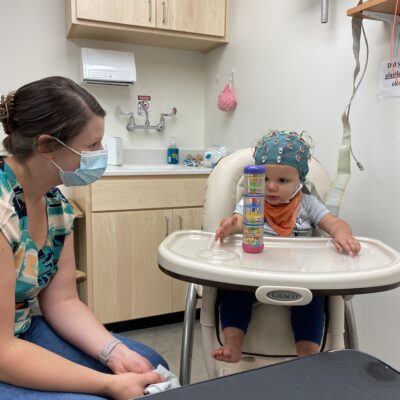 Female lab researcher sitting next to baby boy in high chair wearing teal lab helmet