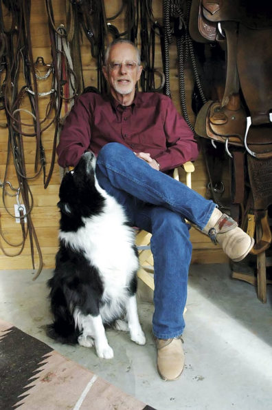 Don Weller seated with his dog at home in front of bridles and saddles