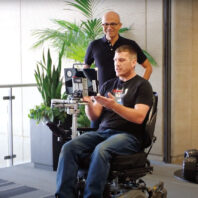 Jon Campbell sits in a wheelchair operated by eye-control. Microsoft CEO Satya Nadella stands behind watching.