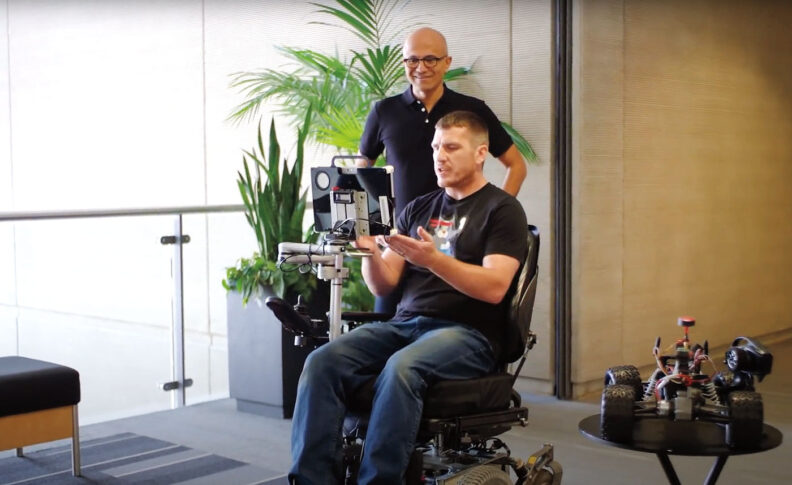 Jon Campbell sits in a wheelchair operated by eye-control. Microsoft CEO Satya Nadella stands behind watching.