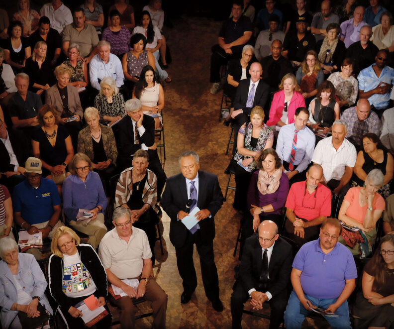 Journalist Enrique Cerna with a microphone and note cards among seated people at a Yakima town hall