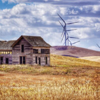 An abandoned wood building in a field with modern windmills behind it