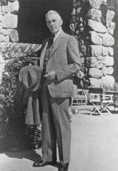 Architect Rudolph Weaver in a suit, holding a fedora, in front of stone walls