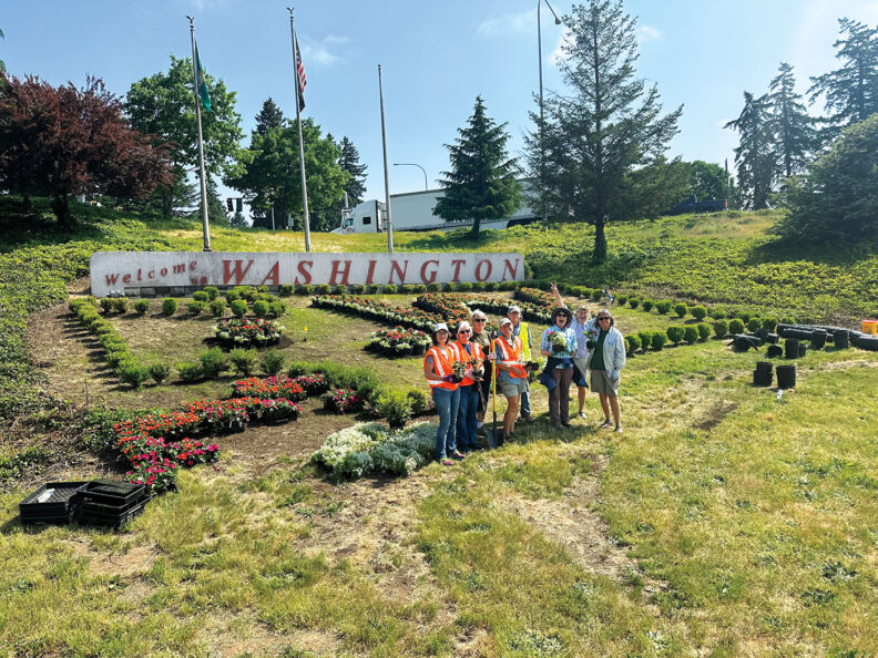 Group of people stand in front of plants and Welcome to Washington sign in Vancouver