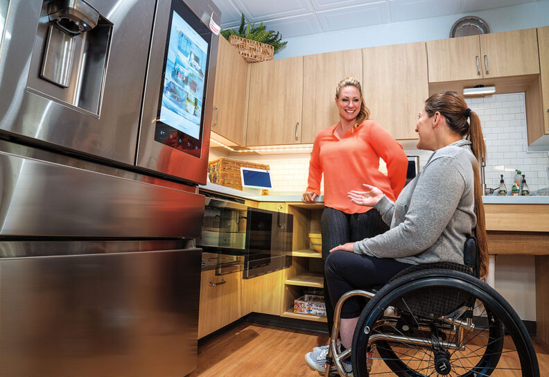 Woman in wheelchair looks at refrigerator while talking to a standing woman