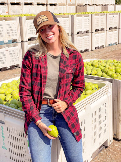 Kaitlyn Thornton in baseball cap, flannel shirt and jeans in front of crates of pears