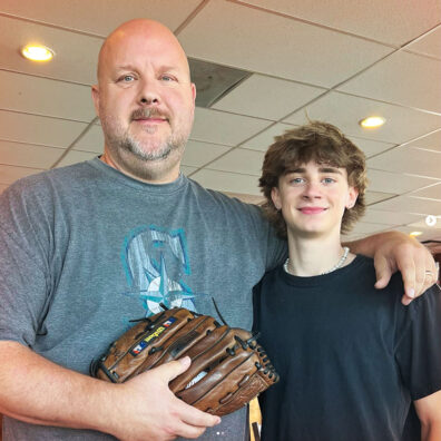 Man in a Seattle Mariners shirt holds a baseball mitt, while standing next to a teen boy