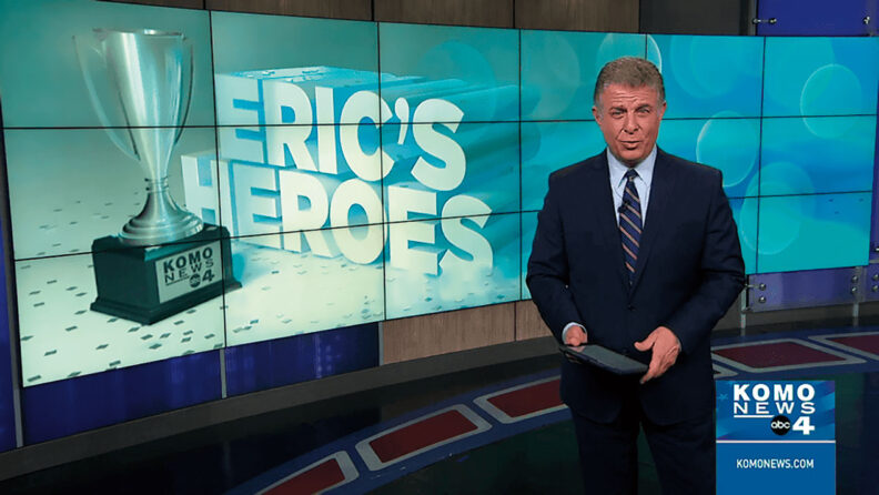 Broadcaster Eric Johnson in front of KOMO 4 News screens showing Eric's Heroes logo