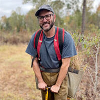 John McGuire near a forest wearing a red safety vest and leaning on a tool