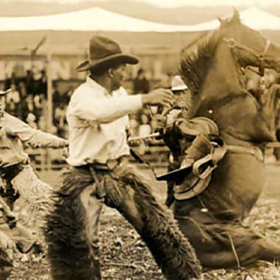 Old photo of a man in cowboy hat and chaps moves away from a falling bronco in a rodeo