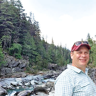 Michael Shepard in a cap stands by a river in a forest