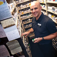 Letters line the wall of a pharmacy as a man, the pharmacist, smiles