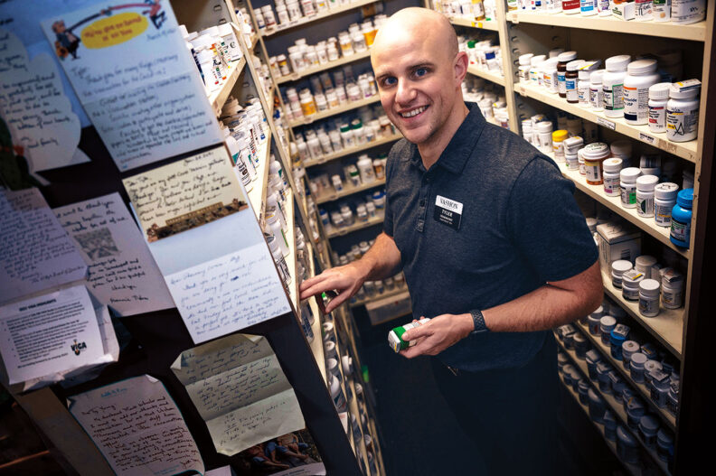 Letters line the wall of a pharmacy as a man, the pharmacist, smiles