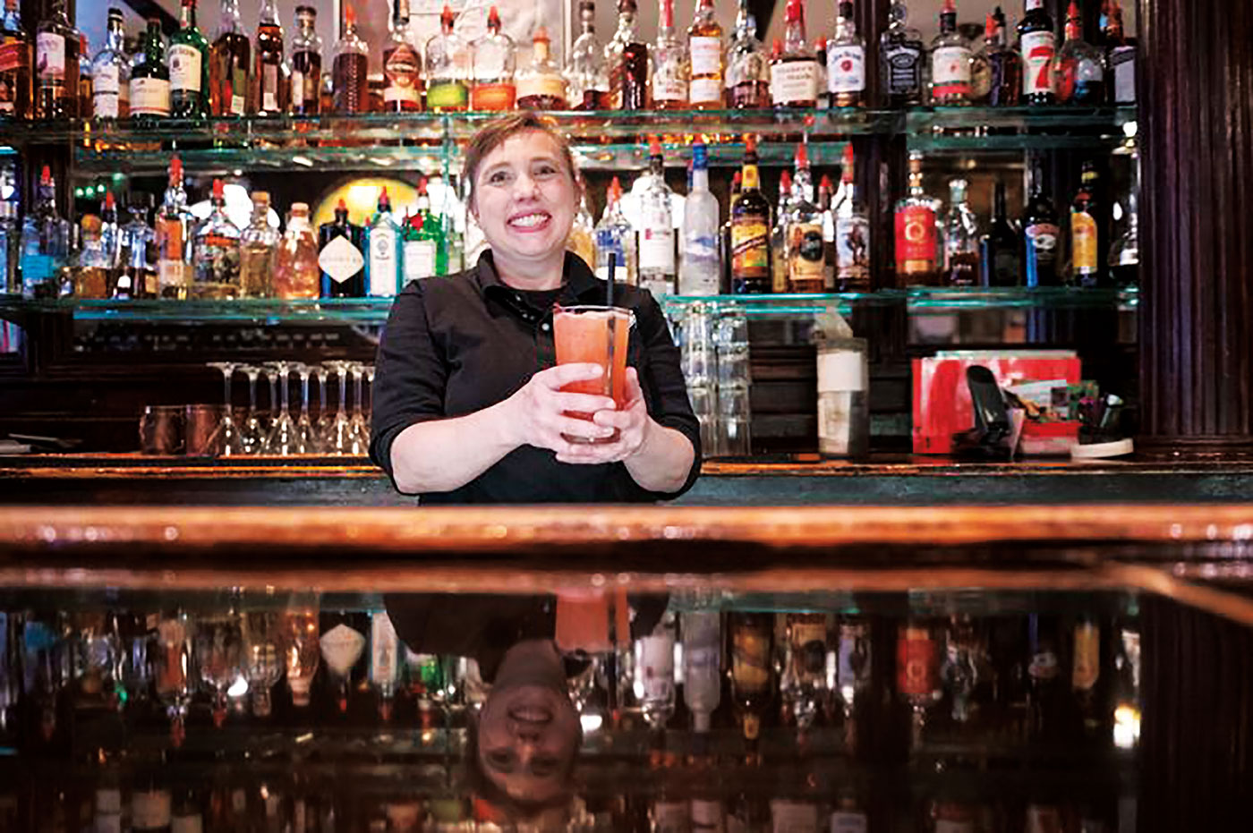 Smiling woman stands behind a bar holding a reddish-orange cocktail