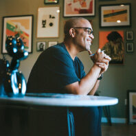 Smiling man seated at a table with framed art on the wall behind him