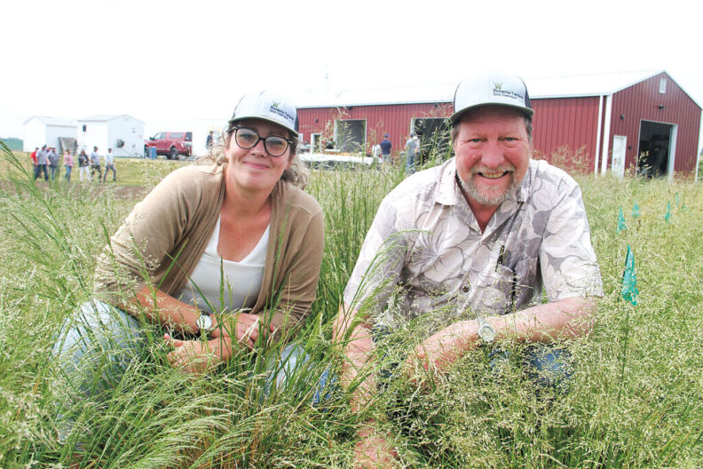 Woman and man crouch in a grass field with metal building behind them
