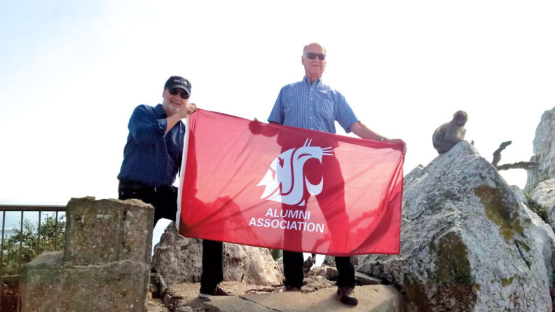 Two men hold a WSU Alumni Association flag at the Rock of Gibralter while a monkey watches