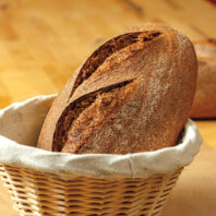 Loaf of bread in a basket in foreground with several other loaves behind it on a table