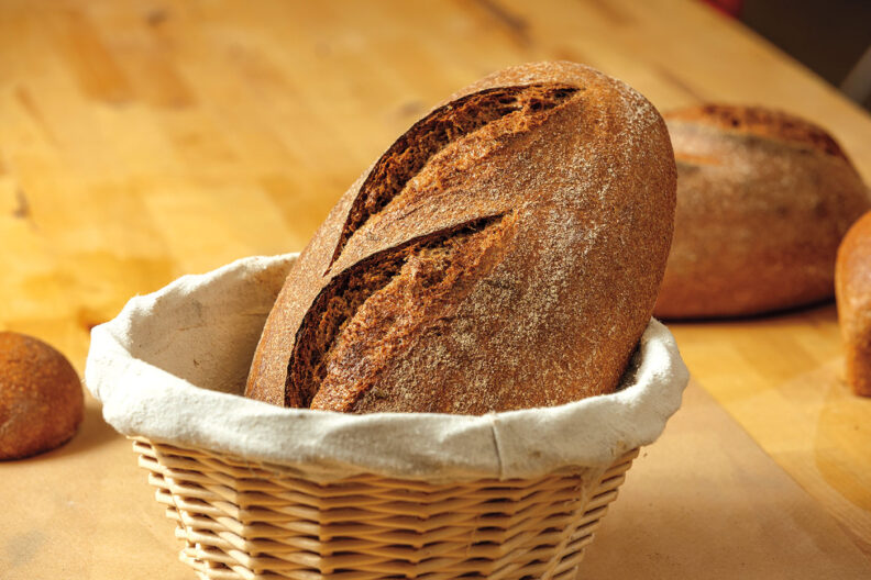 Loaf of bread in a basket in foreground with several other loaves behind it on a table