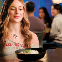 A young woman in a WSU shirt looks at a bowl of gumbo on the table.