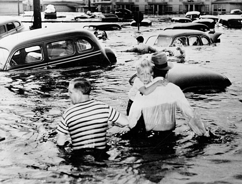 Man carrying a child and another man wading through flood waters as cars float by