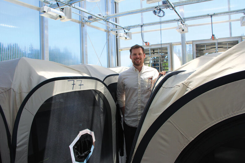 Bearded man stands in a greenhouse lab with large, tent-like cages for fruit flies