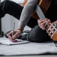 person writing lyrics while holding a guitar in one hand