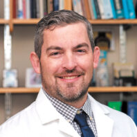 Joshua Neumiller in a lab coat in front of a bookshelf