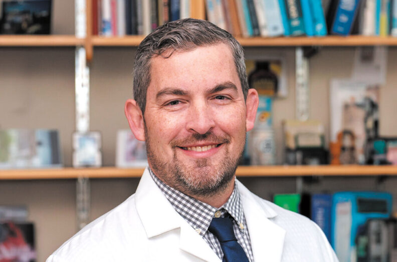 Joshua Neumiller in a lab coat in front of a bookshelf