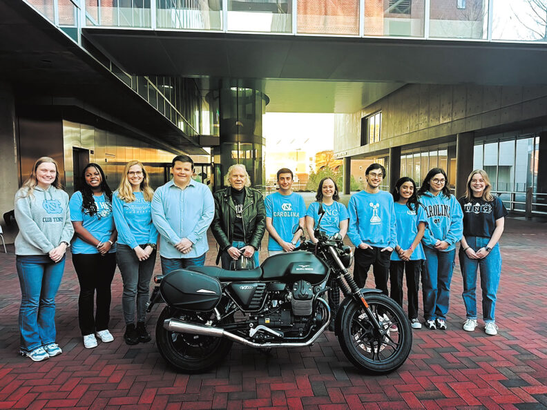 An older man with white hair and leather jackets stands with group of college students behind a motorcycle