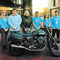 An older man with white hair and leather jackets stands with group of college students behind a motorcycle