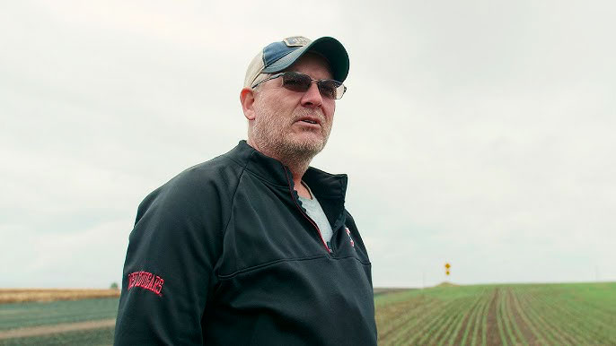 Older man in a farm field, wearing a baseball cap, sunglasses, and WSU Cougars coat