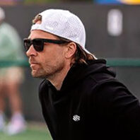 Profile of middle-aged man in sunglasses, black hoodie, and white baseball cap focusing intensely