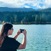 Young woman with long dark hair and glasses takes a picture of a river and forest with her phone