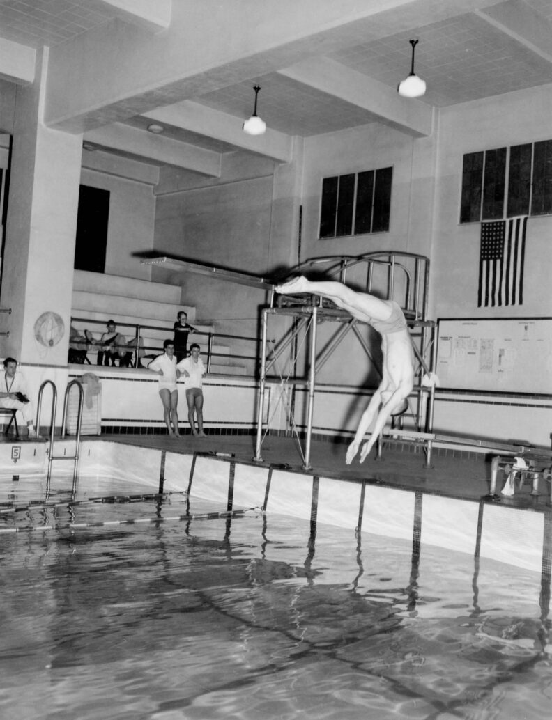 black and white photo of a young man diving into a pool. Several people in the background are watching