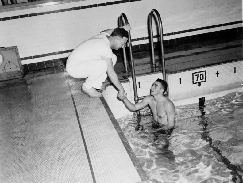Black and white photo of a young male swimmer at the edge of a pool shaking his coach's hand
