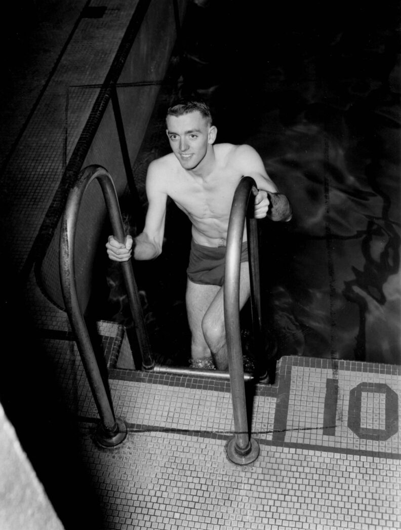 Smiling young man in swim trunks climbs up the ladder out of a pool
