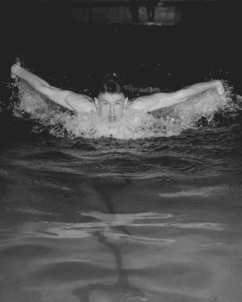 Black and white photo of a young man practicing his butterfly stroke, swimming in a pool.
