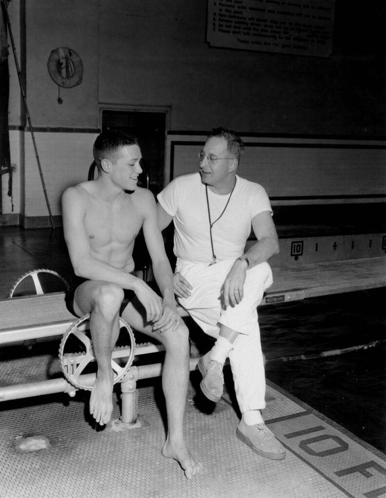 Black and white photo from 1958 of young male swimmer in trunks sitting and talking with older coach in white t-shirt and white pants.