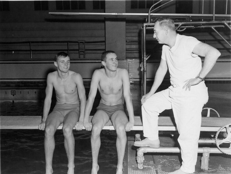 Two young male swimmers in trunks sit on a diving board with coach in white t-shirt and white pants stands next to them.