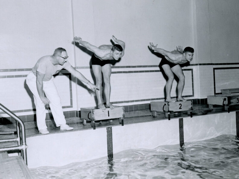 black and white photo of two young male swimmers in trunks preparing to dive into a pool from starting blocks as their older coach points at the water.